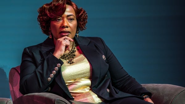 Black woman in black blazer and gold blouse sits in chair on stage during talk