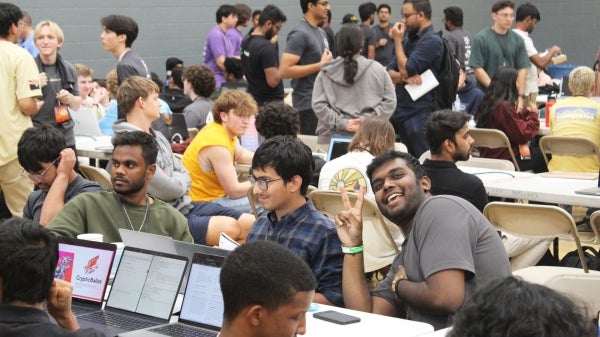 A large group of students sit at tables with laptops and mingle in a large room 