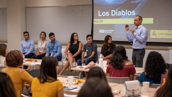 A group of people gather in a room focusing on a man standing in front of a presentation that reads "Los Diablos"