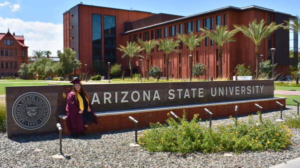 Rhiannon Elliott-McGaugh-Mask sits in front of an Arizona State University sign in her graduation regalia