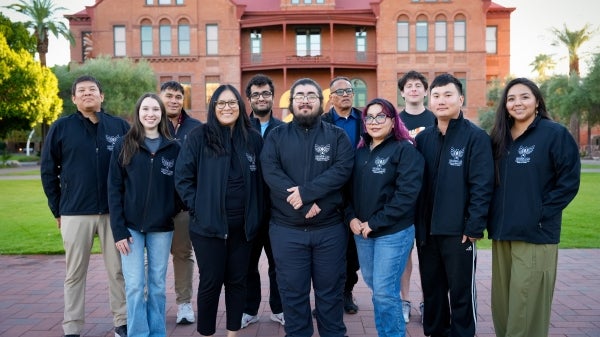 A group of people wearing matching black jackets pose for a photo in front of ASU's Old Main building.