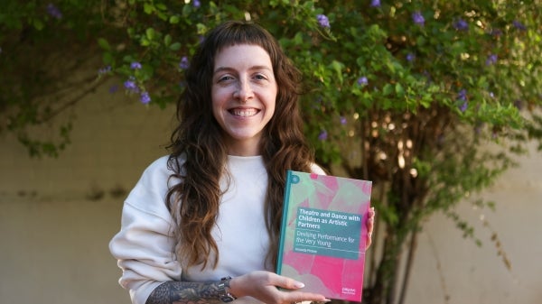 A woman stands under a tree holding her book with teal and green cover.