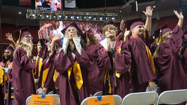 People in maroon caps and gowns wave, smile and blow kisses to an unseen audience in an arena.