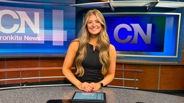 A young woman with long blonde hair wearing a black sleevless dress sits behind a news desk