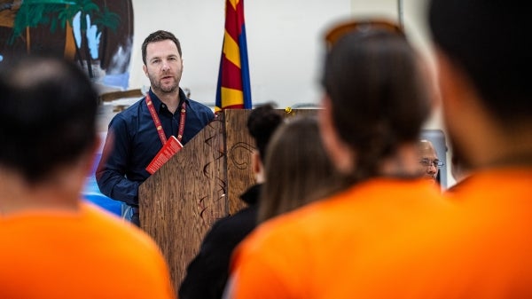 Man speaking behind lectern to group of male inmates wearing orange jumpsuits