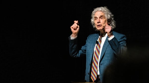 White man with gray curly hair wearing a suit and tie raises his hands to make a point while giving a lecture