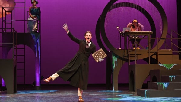 A student gestures center stage with a broad grin while in the background a student sits on an elevated stage to the left and a musician on an elevated stage to the right.