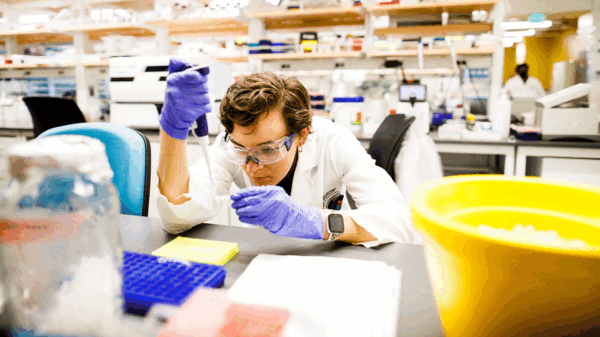 Student in lab gear hunches over lab table to fill vials with a pipette