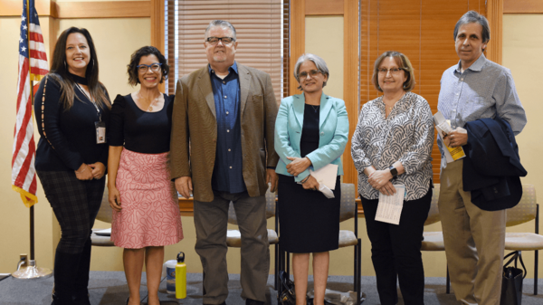 Six language and translation professionals stand next to each other on a stage. They are wearing business casual clothing. There is an American flag in the background.