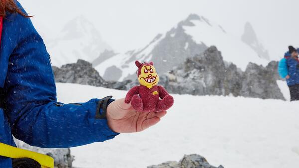 Hand holding a stuffed Sparky the Sun Devil against a backdrop of a snowy, mountainous landscape.