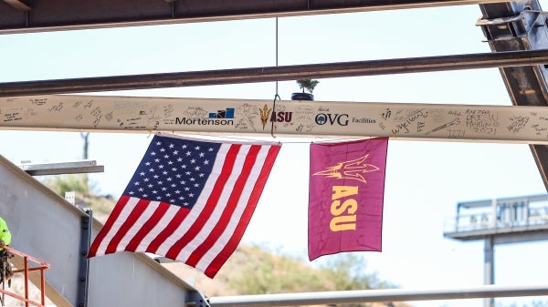 A steel beam covered in signatures and attached to two flags is being lifted by a crane