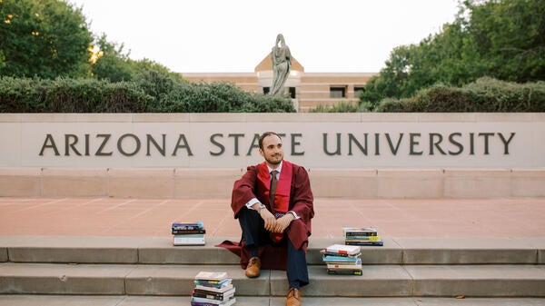 Bryson Brown sitting on steps in front of Arizona State University in graduation gown surrounded by books