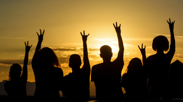 Silhouettes of a group of people in front of a sunset holding their hands in the air in the shape of an ASU pitchfork.