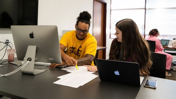 Two college-age people sit at a computer desk looking at papers