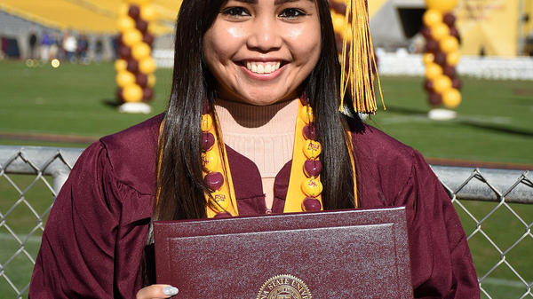 Danni Baquing smiling and posing with ASU degree wearing graduation gown and stole in an outdoor setting with maroon and gold balloons behind her.