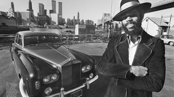 Black-and-white image of a man in a coat and hat in front of a classic car.