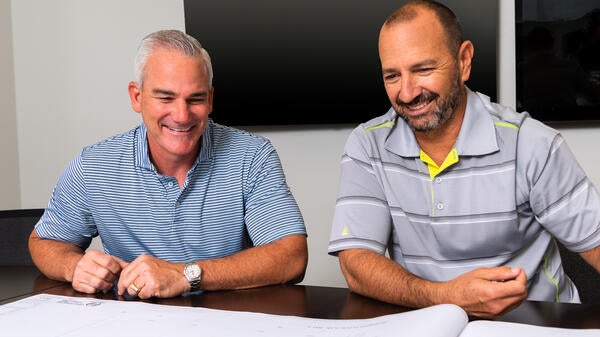 Two men sit side by side at a table looking down and smiling.