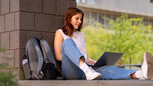 A person sitting with their back against a pillar as they're on their laptop.