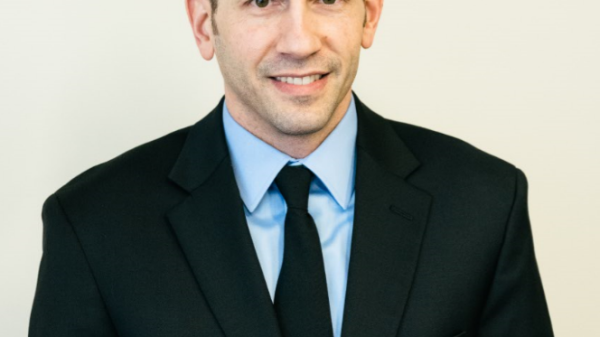 A brunette man in a black suit smiles at the camera.