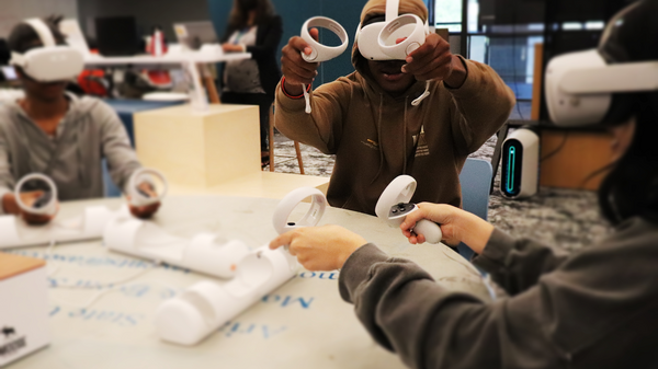 Students sit around a table wearing virtual reality headsets and handheld controllers