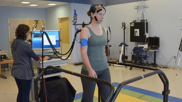 A woman with a sensor device on her head walks on a treadmill while a researcher monitors readings on a computer