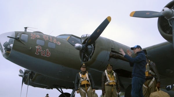 Matthew Wilcox stands in a dark blue hoodie with three actors dressed in military air uniforms next to a retired military plane.