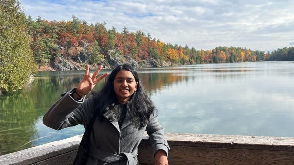 Anusha Natarajan holding up the ASU pitchfork hand sign in Canada, standing on a deck in front of a lake surrounded by trees.