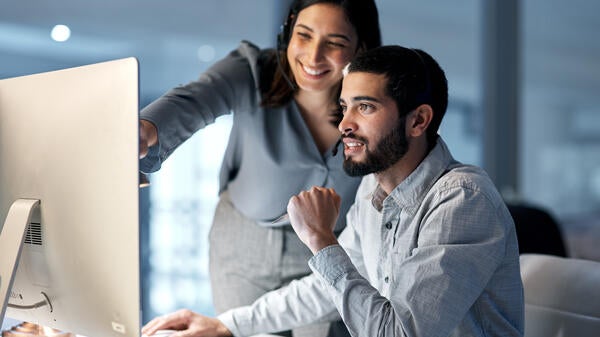 Woman standing next to a man seated at a computer. They are both smiling as the woman points at the computer screen.