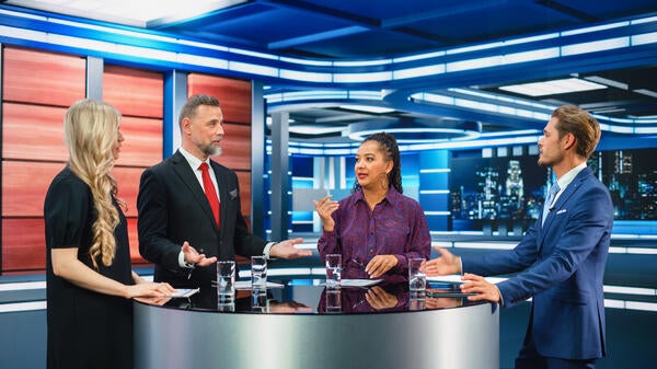 Four broadcast  journalists stand around a news desk talking