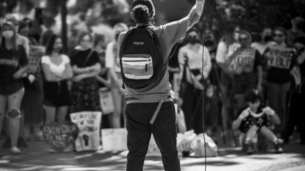 Black-and-white photo of a person seen from behind speaking into a microphone to a crowd of people.