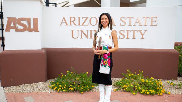 Kristin Payestewa-Picazo stands in front of an Arizona State University sign. She is wearing traditional clothing representing her Hopi and Navajo heritage. She has a feather in between her hands and smiles at the camera.