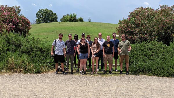 A group of students posing in front of a hill surrounded by greenery.