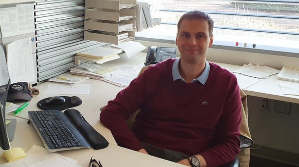 Petr Šulc smiling at the camera and sitting at an office desk with a computer and papers surrounding him.