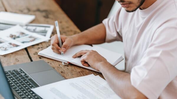 Person seating at a table in front of a laptop writing in a notebook.