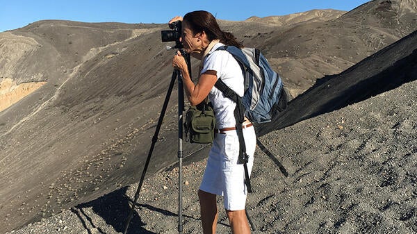 A dark-haired woman in white shorts and a white t shirt stands at a tripod on a gravel hill, taking a photograph