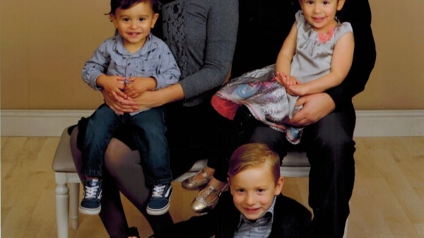 Rachel Hill, wearing a gray sweater and vivid pink lipstick, sits next to the right of her husband. They are each holding one of their children on their laps, and a third sits on the floor in front of them. They are posing for a family portrait.