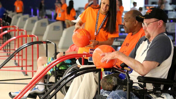 A woman holding a bowling ball speaks to two men in wheelchairs holding bowling balls.