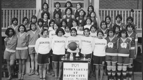 Black-and-white photo of a group of Native American students posed for an athletics photo on a staircase at a school.