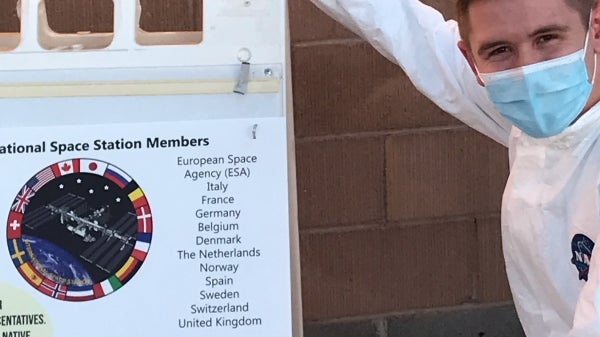 Eli Fox squats in front of a brick wall. He is wearing an all-white jumpsuit with a blue NASA logo over the breast. He has short brown hair and is wearing a blue surgical mask. His arm is extended to point at a sandwich board displaying scientific facts.