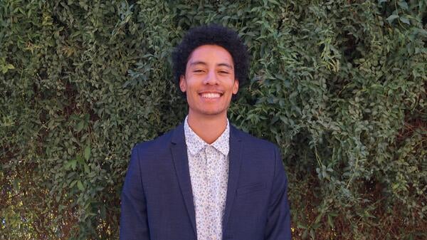 Man wearing collared shirt and blazer smiles in front of wall of greenery