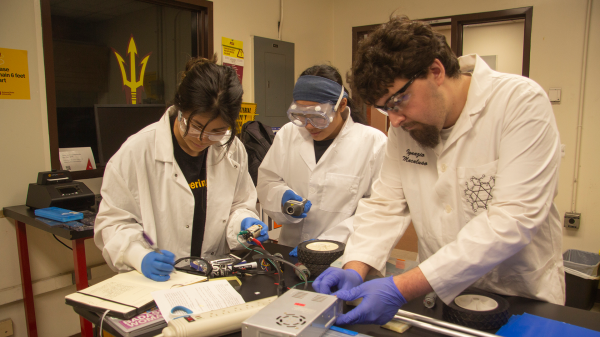 Three students in lab gear examine their car battery using various instruments.