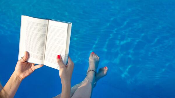 Hands holding a book above a pool in which a person's legs are dipped.