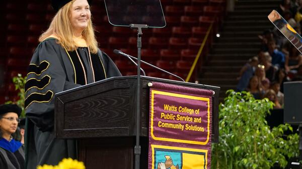 Woman wearing graduation regalia, standing behind a lectern.