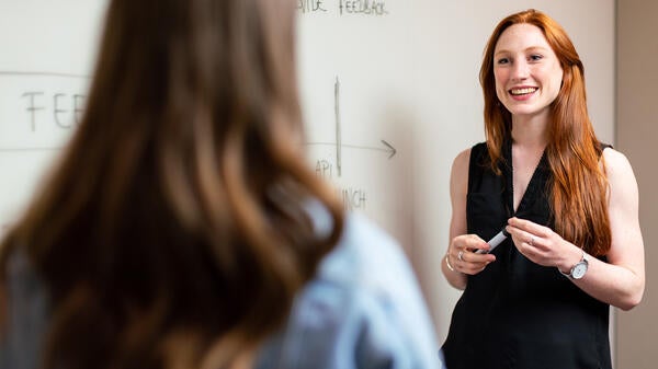 Woman standing next to a whiteboard and smiling while looking at another woman facing her, seen from behind.