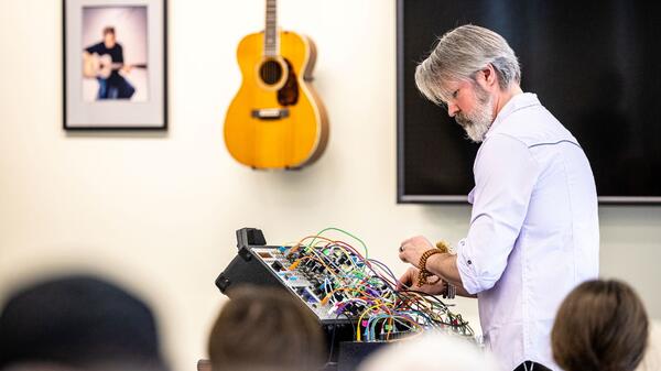 Man standing at a board with several wires poking out of it in front of a wall with a guitar hanging on it.