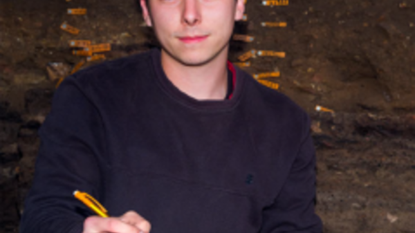 Patrick Fahey sitting down holding a pencil and writing in a notebook while looking at the camera.