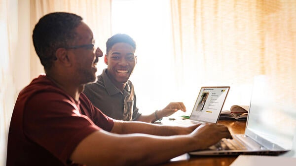 Two Black men smile as they work at laptops.