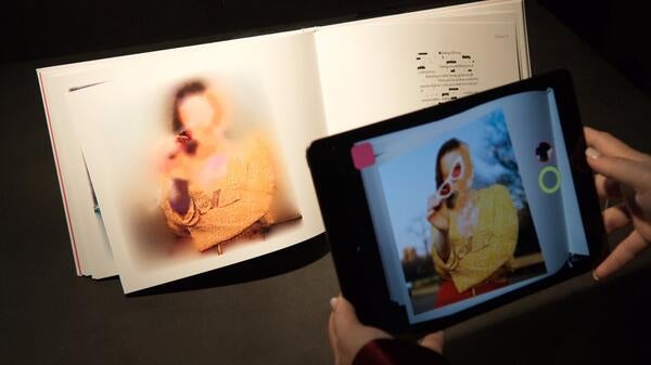 Hands holding a device up in front of a book. The book shows a photo of a faceless person while the device displays a person with a face.