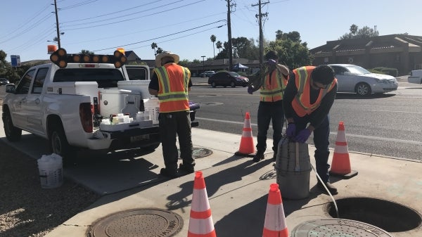 Crew of workers wearing bright orange vests oversee a tank attached to a hose that has been inserted into a city manhole.