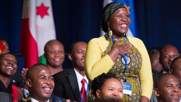 A young African woman stands among peers with her hand over her heart. 